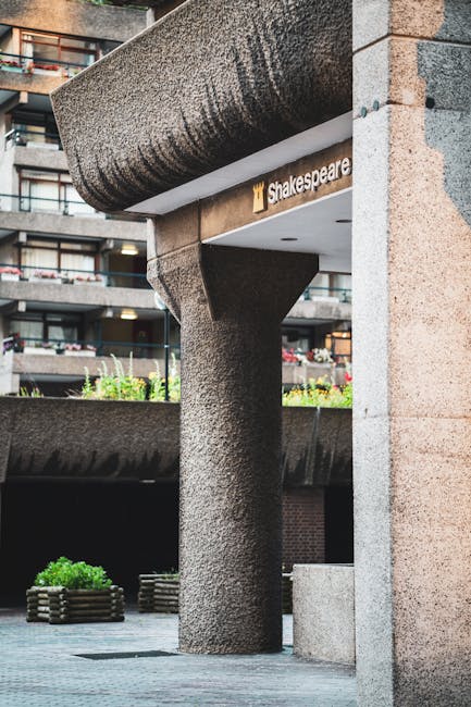 A close-up view of a modern urban building's exterior, featuring a textured, cylindrical concrete support column with a curved capital, supporting a large, rounded overhang made of similar rough-textu